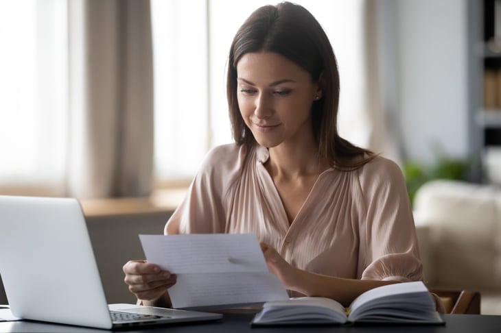 Happy woman smiling and reading a printed page that might be a credit report or legal document while sitting at her laptop