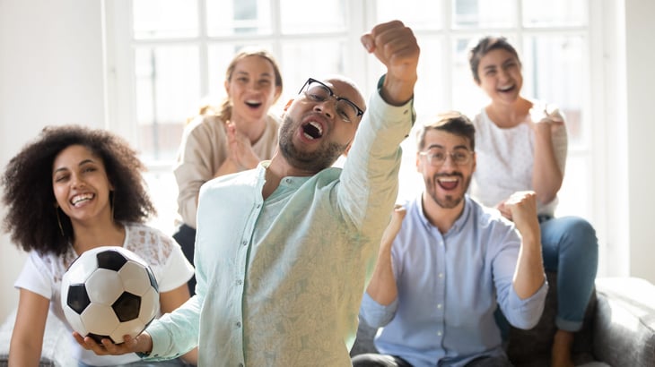 Friends excited and watching a sports game with a soccer ball and cheering in front of the TV