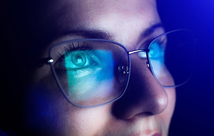 Woman wearing glasses staring at a computer with a closeup on her eyes looking at monitor full of blue light