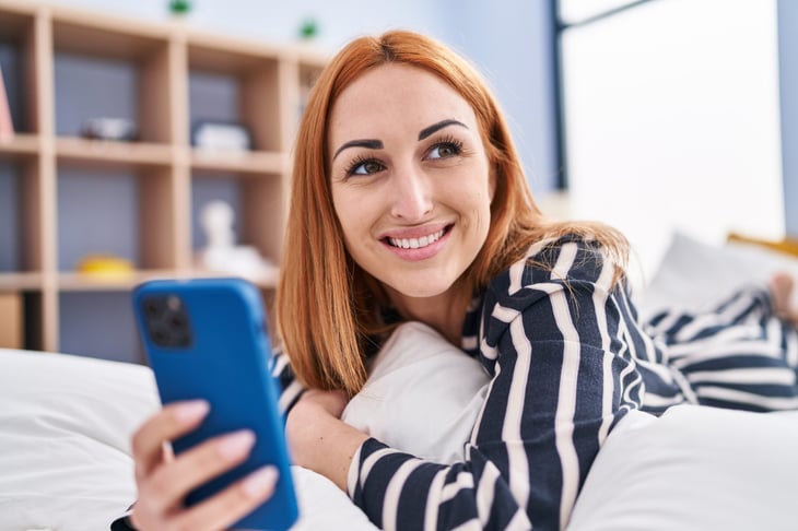 Happy woman smiling and laying on the bed with her smartphone taking pictures or browsing the internet
