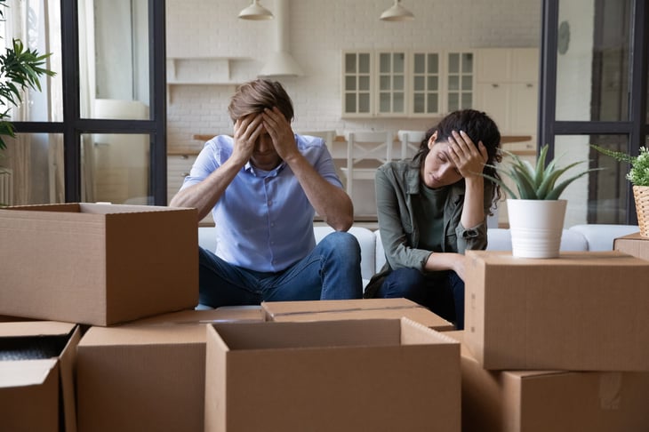 Exhausted young couple sit rest on sofa in living room near heap of cardboard boxes