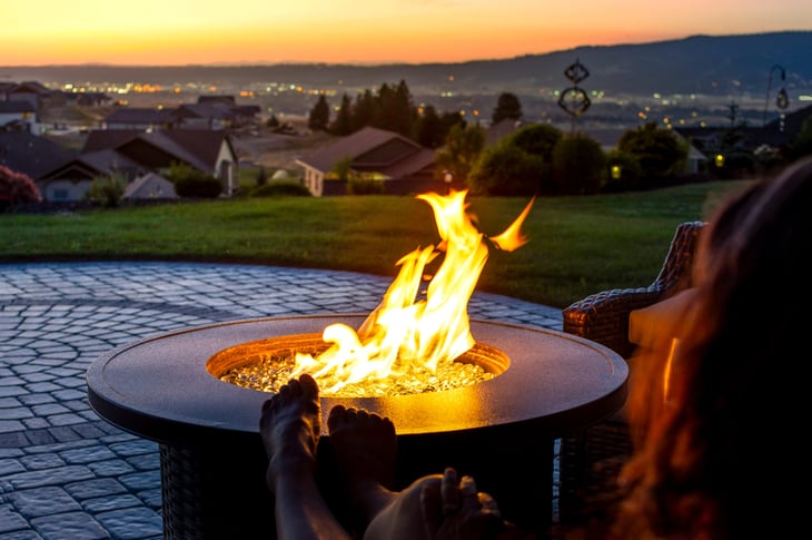 A woman relaxes by a roaring firepit on a paver patio at sunset