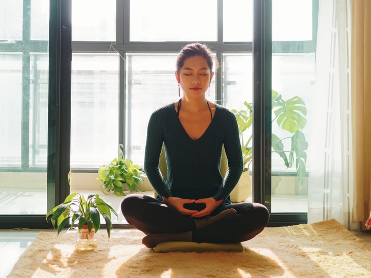 Relaxed woman doing meditation and yoga in a well lit room full of sunlight and plants to remove stress