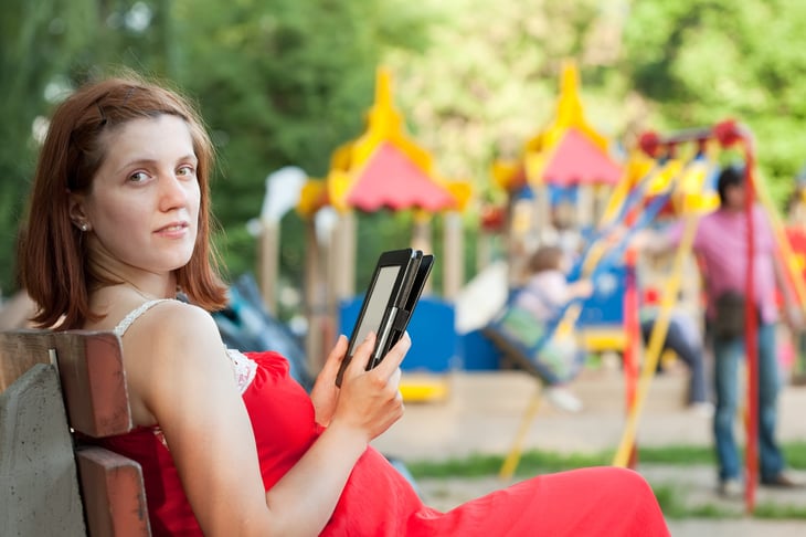 Pregnant woman sitting on a bench in a park at the playground watching children and reading an e-reader or tablet