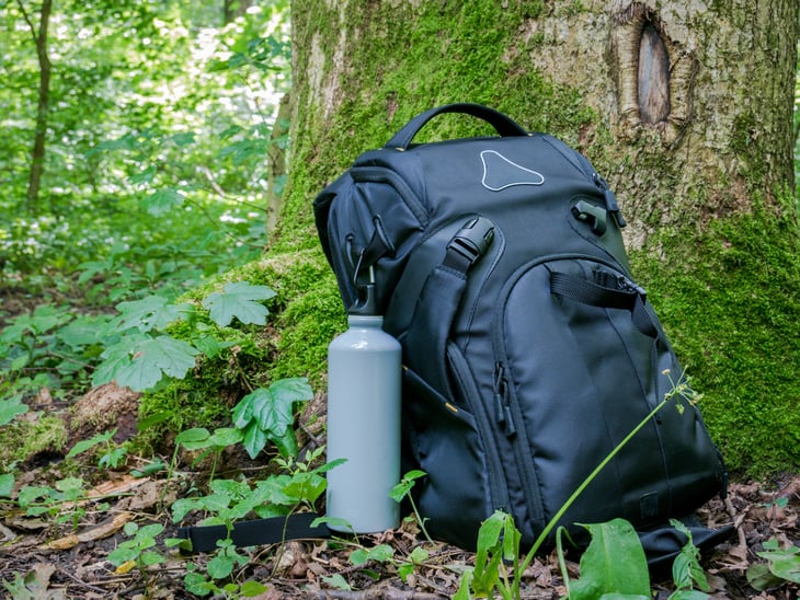 A backpack and reusable water bottle next to a tree trunk with green moss