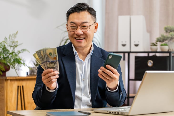 Happy rich guy with a handful of cash, holding a phone in front of a laptop