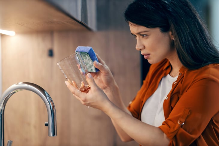 Woman squinting at dirty dishes and holding a soapy sponge up to a glass in the kitchen sink area
