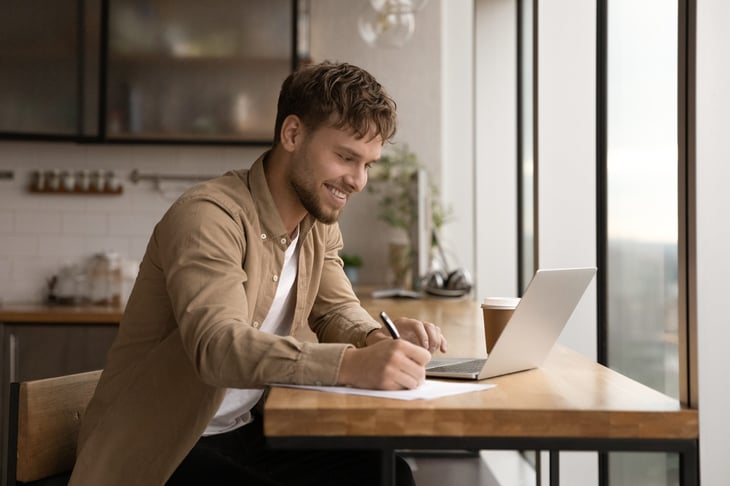 A young man smiles while sitting at his laptop and taking notes