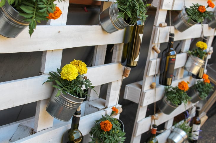 Metallic pots with yellow, orange flowers hang on a white pallet fence.