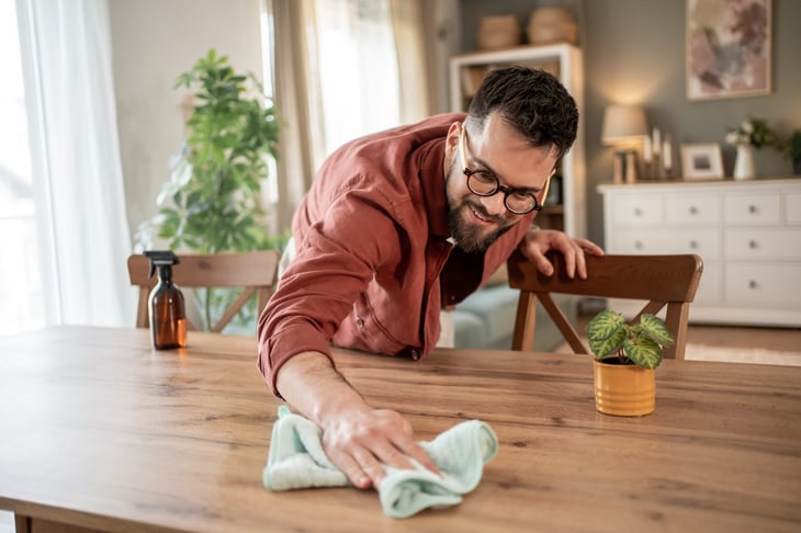 Man wearing glasses is carefully cleaning a wooden dining table with a microfiber cloth.