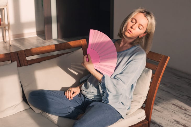 Hot and sweaty woman on couch overheating and using a fan to cool herself
