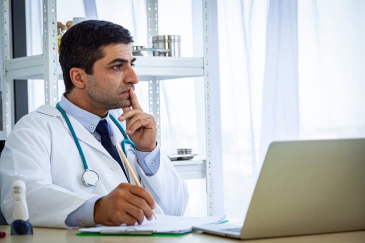 Doctor looking thoughtful using a laptop and writing down information