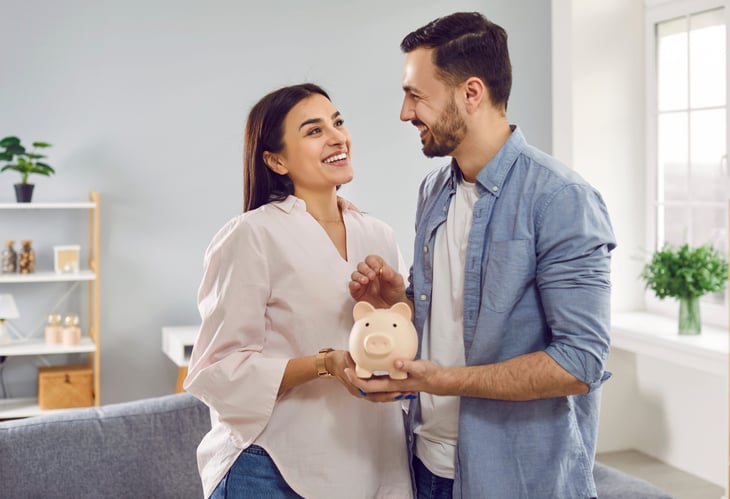 A couple smiles while holding a piggy bank and putting money into it.