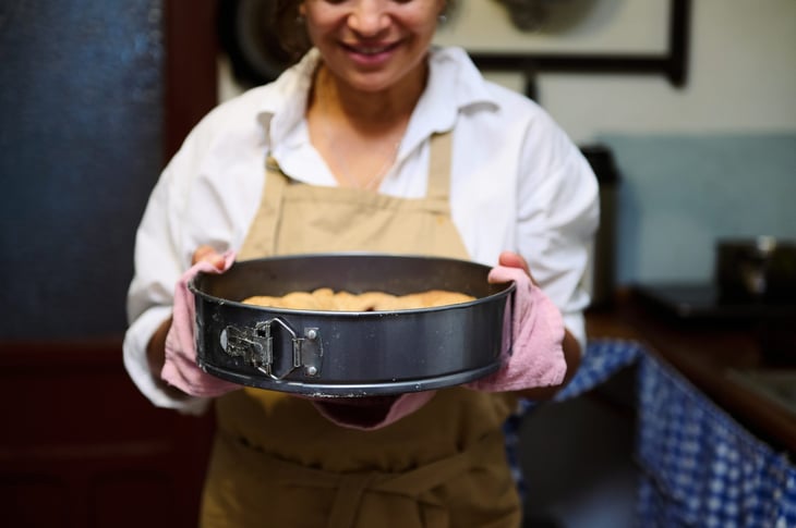 A smiling woman wearing an apron and oven mitts holds a freshly baked cake in a springform pan, freshly out of the oven