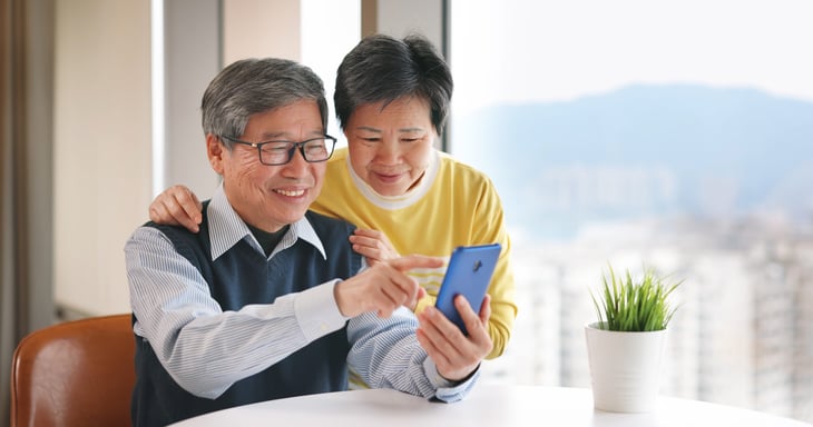 Senior couple smiling and looking happy while pointing at smartphone in front of a bright window at home