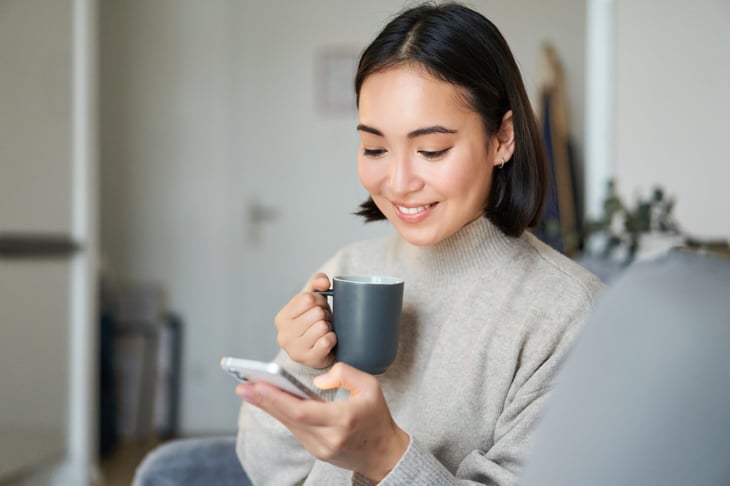 Happy woman sipping coffee and wearing a sweater while smiling at her phone
