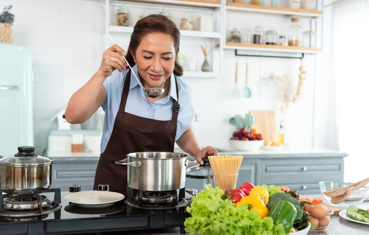 Smiling woman cooking over a stovetop, about to take a sip from a ladle