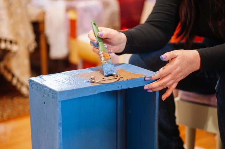 Woman painting an antique dresser drawer bright blue with a paintbrush to restore the furniture