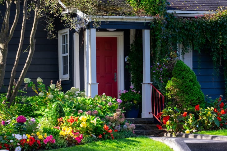 A red door on a wooden navy blue house with white trim. The cottage has lush greenery around the entrance: red, yellow, pink, and white flowers, hydrangea, and green grass.