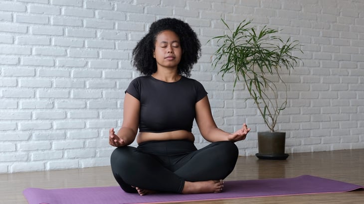 A woman does yoga and meditates on a mat at home.
