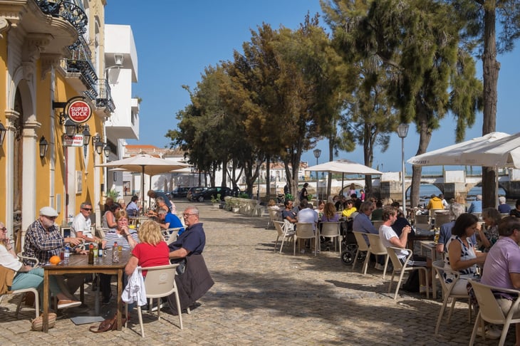 Outdoor cafes along the Rio Gilao river in Tavira, Portugal, where people eat and drink on a sunny spring day.