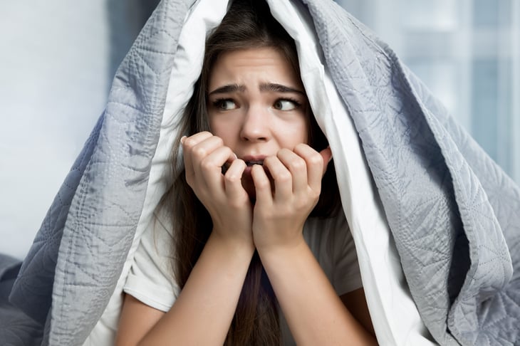 Scared young woman hiding under the covers in bed.