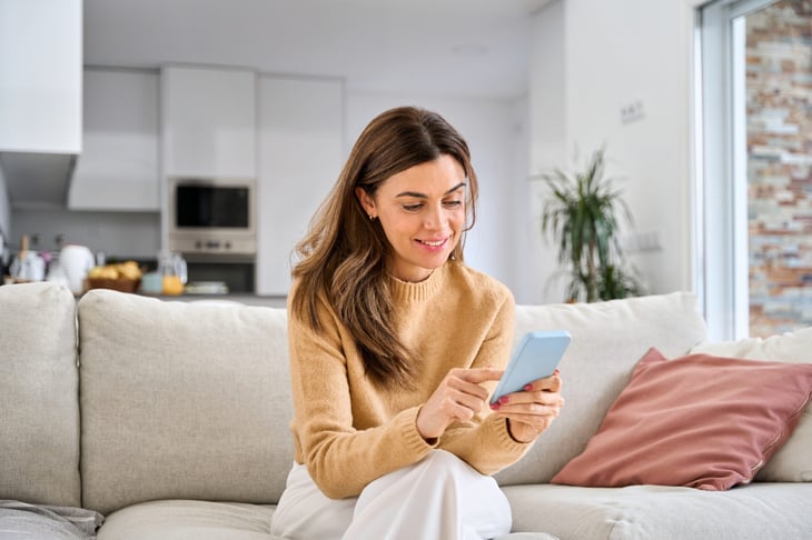 Middle-aged woman smiling as she sits on a couch and uses her smartphone.