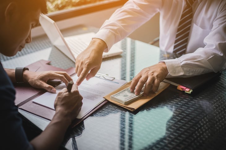 Man signing documents for a loan or payment of a debt and another pointing to the signature line and holding a pile of cash