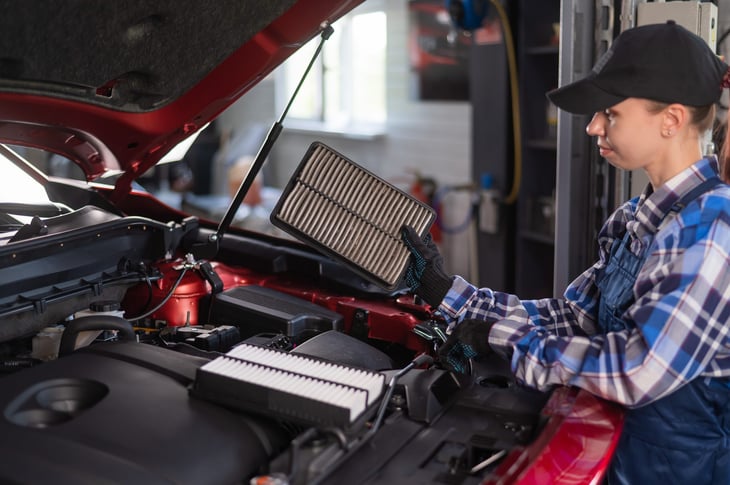 Woman mechanic replacing an engine air filter under the hood of a car in a garage or auto shop