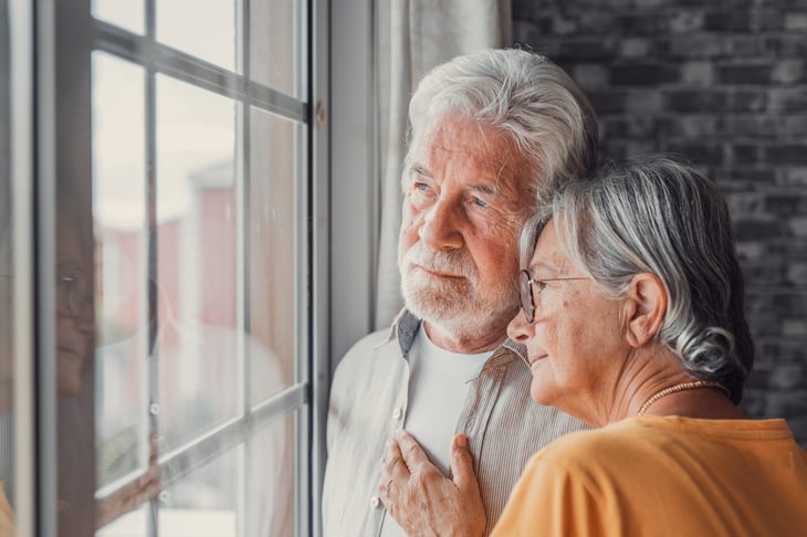 Pensive senior man looking out a window. Old woman consoling and hugging him.