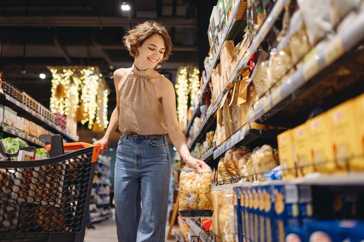 Smiling young woman picking up a package of pasta in a grocery store, pushing a cart.