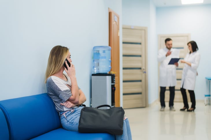 Woman talking on her smartphone in a hospital waiting room with two doctors in the background.