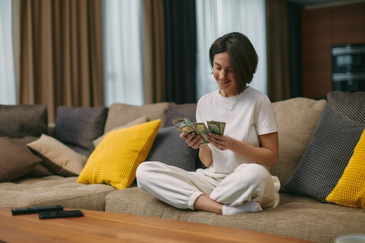 Happy young woman counting cash, sitting on a sofa at home