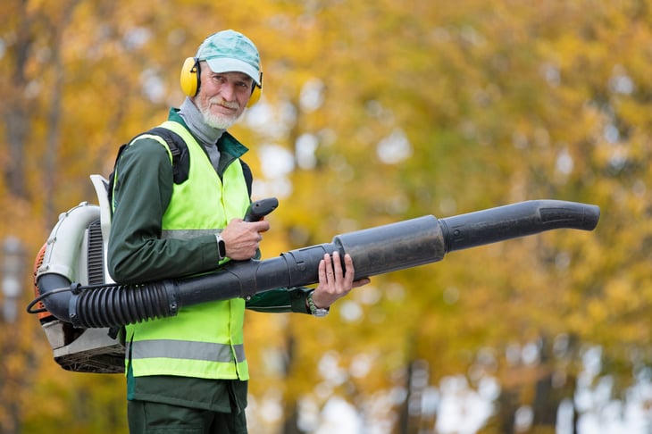 Smiling senior man cleaning the yard with a leaf blower in autumn outdoors
