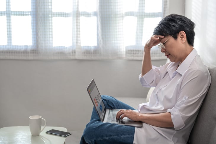 An older woman is stressed while sitting on a couch, looking at her laptop
