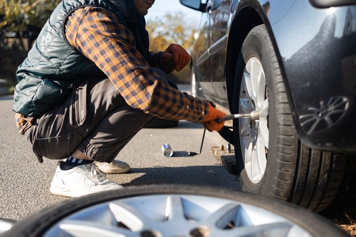 A man wearing work gloves changes a tire on a car on the side of a road