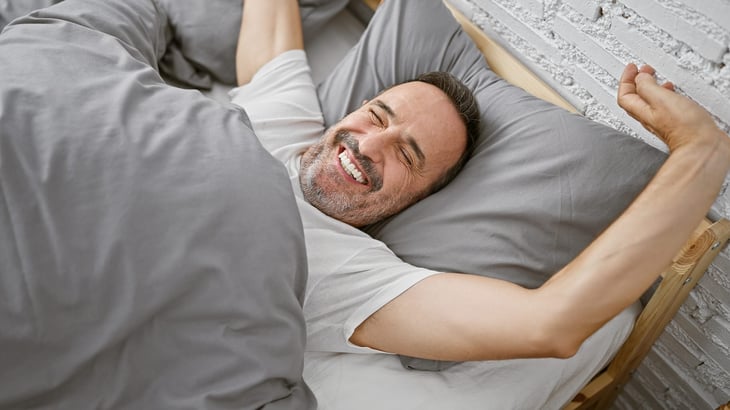 Smiling middle-aged man in bed, waking up happy and well-rested after a good night's sleep.