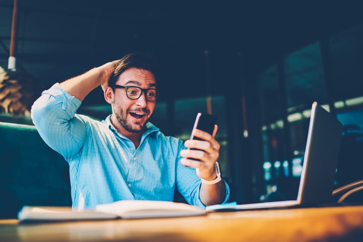 A surprised and excited man with a laptop looking at his cell phone.