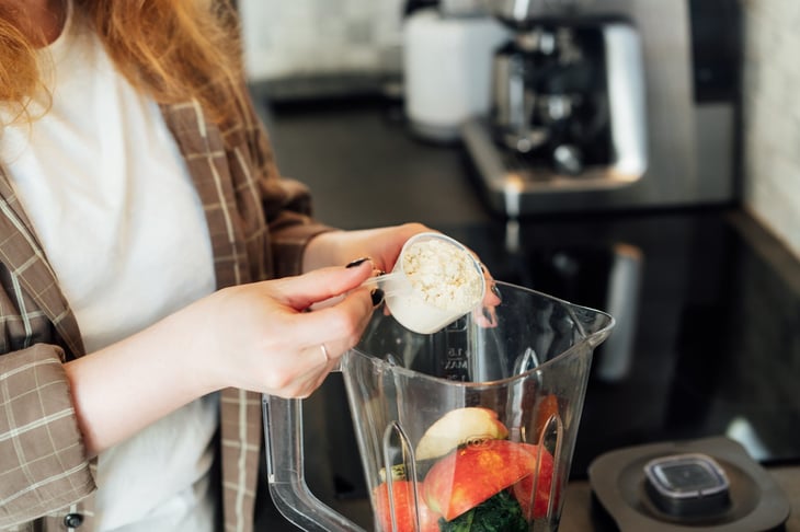 Woman adding a scoop of collagen or protein powder to a blender full of fruits and vegetables for a healthy smoothie.