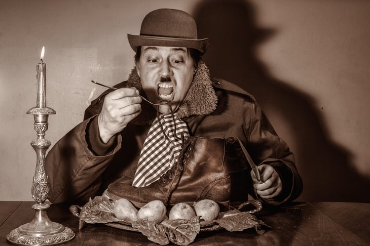 Poverty-stricken, bankrupt, poor male with mustache eating a boot by candle light in a dirty, cold room, wearing a hat.