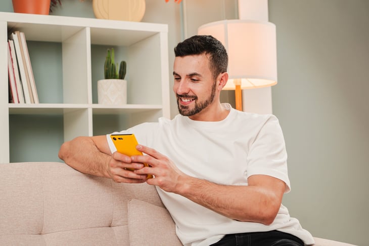 Young Man Sitting Comfortably on Couch Using Smartphone with a Bright Yellow Case