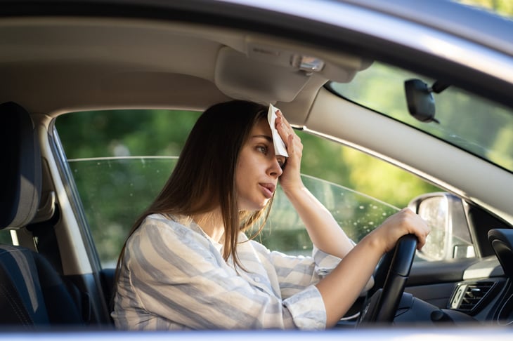 Woman sweating in a car that is too hot in the summer and overheating with a wash cloth on her forehead to cool off