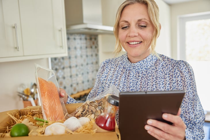Woman holding a tablet and unpacking a meal kit delivery.