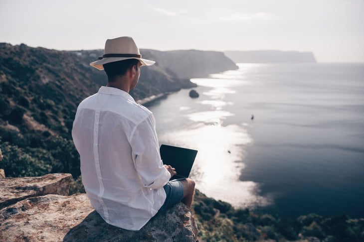 Digital nomad works remotely on a laptop while sitting on a cliff overlooking a beach