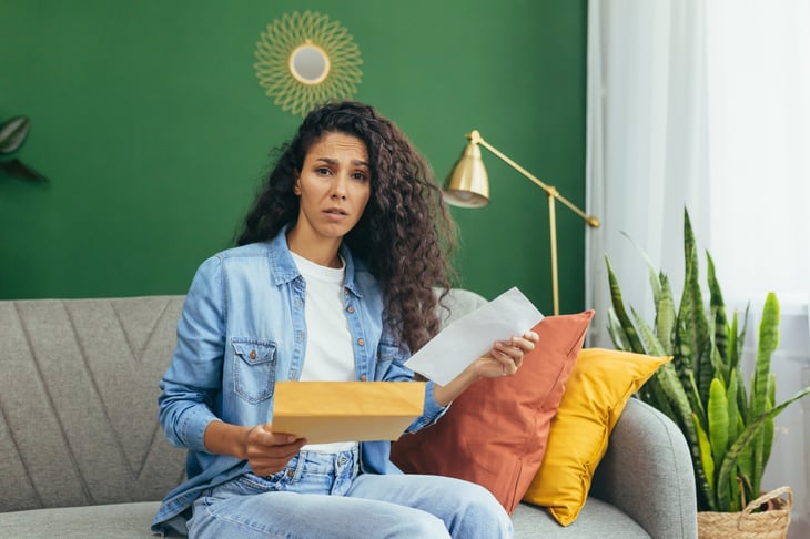 Disappointed and sad woman sitting on a couch at home holding a paper and envelope.