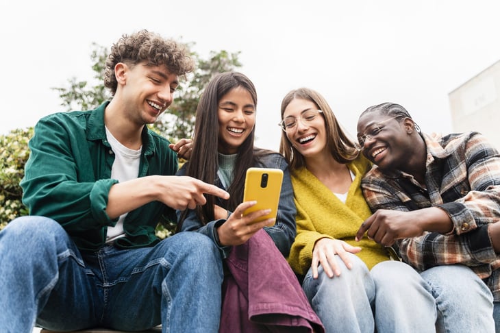 Group of young Gen Z friends sitting together using cell phone app to share funny social media content