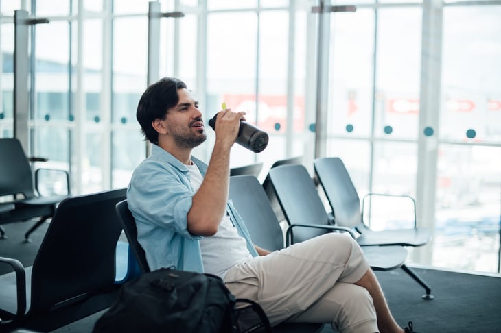 A young man waiting at the airport gate to board a plane, with a backpack and drinking from a water bottle