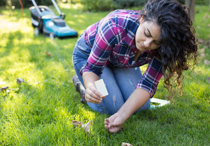 A woman measures grass height for trimming. Mower in background.