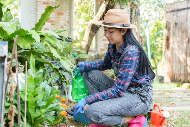 woman in hat and plaid shirt, kneeling and spraying plants with green bottle in garden.