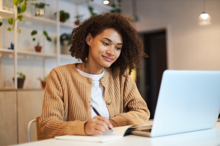 Confident young woman using a laptop and taking notes to write a resume.